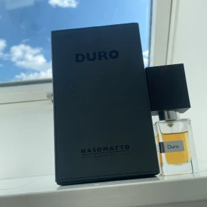 Duro parfym från Nasomatto - Säljer en exklusiv parfym från Nasomatto, Duro. Flaskan har en elegant design med en fyrkantig, mörk träkork och en klar glasflaska som visar den gyllene vätskan. Perfekt för den som söker en unik och kraftfull doftupplevelse.
