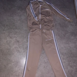 Beige Dress från Palm Angels - Säljer en beige dres från Palm Angels med vita och svarta ränder längs ärmarna. Dressen har en dragkedja framtill och matchande byxor med samma design. Perfekt för varma eller soliga dagar!