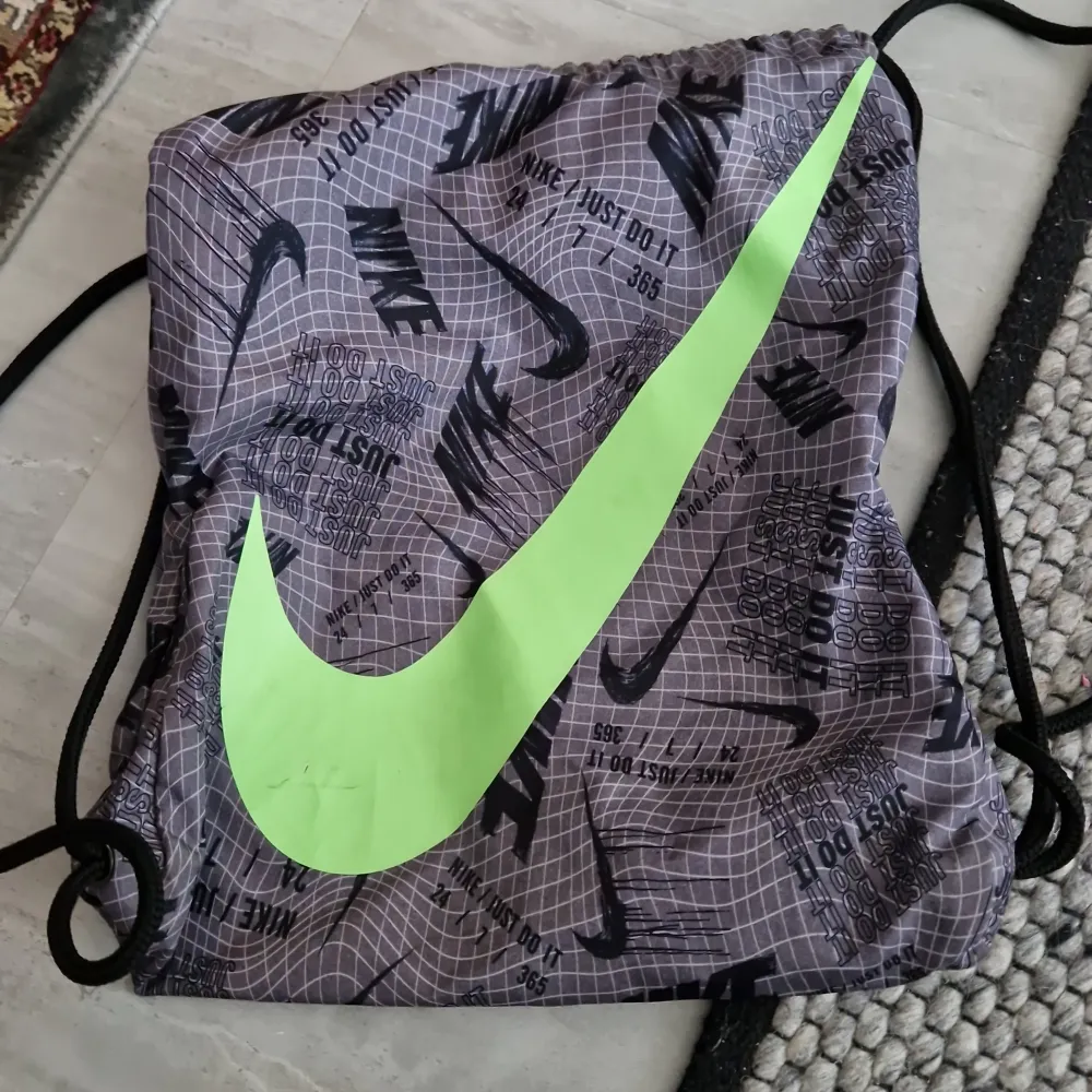 Säljer en grå och svart gympåse från Nike med ett stort grönt swoosh-märke. Väskan har ett mönster med texten 'Just Do It' och Nike-loggor. Perfekt för träning eller som en lätt vardagsväska. OBS! LITET MÄRKE PÅ NIKE MÄRKET!. Laukut & Käsilaukut.