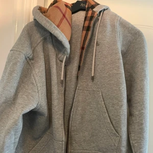 Burberry hoodie - Säljer en stilren grå hoodie från Burberry med dragkedja och ett snyggt rutigt foder i huvan. 