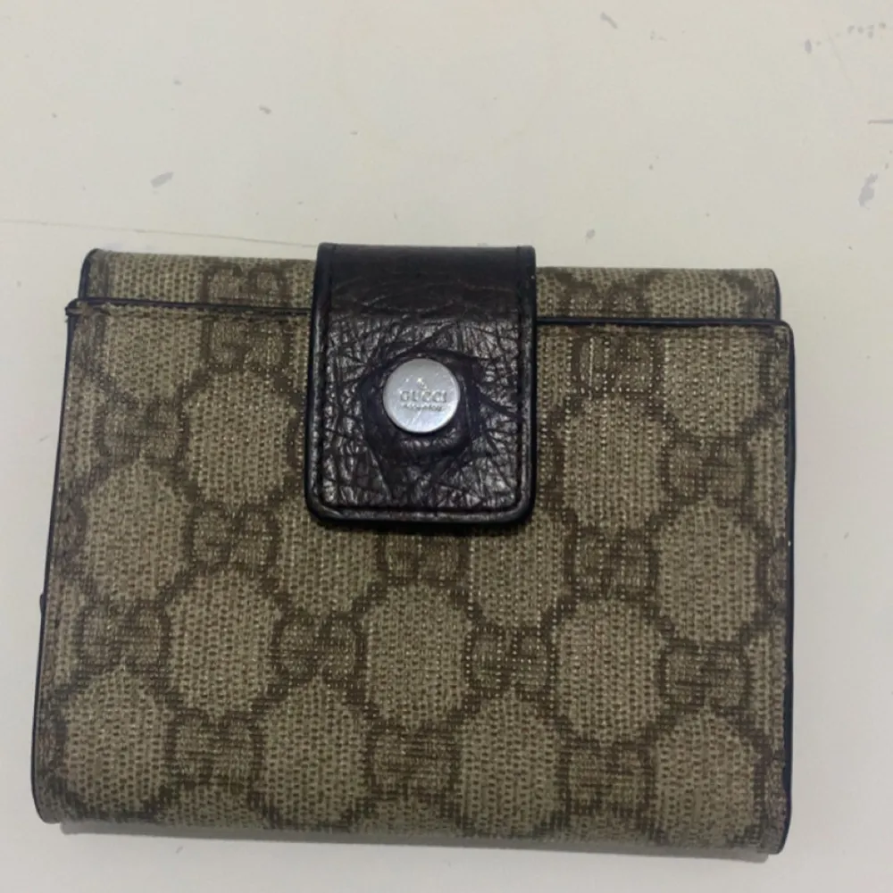 Snygg plånbok i beige och brunt med ett klassiskt mönster. Den har flera fack för kort och en knappstängning i mörkt skinn. Perfekt för att hålla ordning på dina kort och kontanter.. Laukut & Käsilaukut.