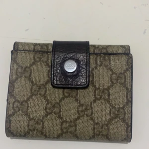 Beige och brun plånbok med mönster - Snygg plånbok i beige och brunt med ett klassiskt mönster. Den har flera fack för kort och en knappstängning i mörkt skinn. Perfekt för att hålla ordning på dina kort och kontanter.