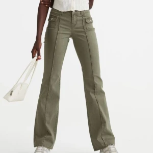Olivgröna bootcut byxor med fickdetaljer - Snygga olivgröna byxor i bootcut-stil med medelhög  midja. Byxorna har fickor med knappar framtill och en dragkedja. 