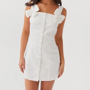 Vit kortklänning med rosetter - Säljer en söt vit kortklänning med knappar framtill och stora rosetter på axelbanden. Supersöt men säljer då den inte passar