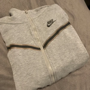 Grå hoodie från Nike - Säljer en grå hoodie från Nike med dragkedja och svart logga på bröstet. Den har en svart randdetalj över bröstet och är tillverkad i mjukt material. Perfekt för en avslappnad stil. Har två sydda hål på framsidan med dom syns knappt.
