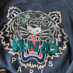 Blå sweatshirt med tigerbroderi från Kenzo - Säljer en blå sweatshirt från Kenzo med ett stort, färgglatt tigerbroderi på framsidan. Tröjan har långa ärmar och ribbade muddar vid ärmslut och nederkant. Perfekt för en avslappnad stil med en touch av lyx. Tröjan har fått klorin på armen därav priset 