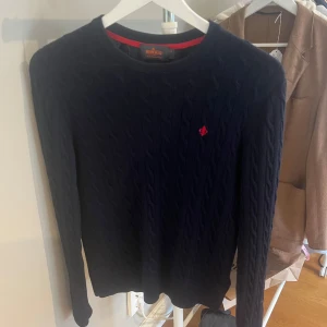 Svart kabelstickad tröja från Gant - Säljer en stilren svart kabelstickad tröja från Morris med ett rött broderat emblem på bröstet. Tröjan har långa ärmar och en rund halsringning. Perfekt för kyligare dagar. Passar XS