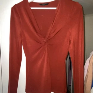 Rostfärgad topp från Vero Moda - Säljer en snygg rödbrunfärgad topp från Vero Moda med en unik knutdetalj framtill. Toppen har långa ärmar och en elegant v-ringning.                                        , använd typ 3ggr. Ingen aning om vad den kostade men gissar runt 2-300. Säljer för 60 iaf   