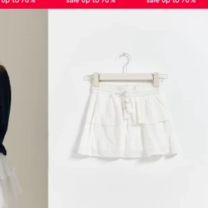 Vit kortkjol med volang och knytband - Söt vit kortkjol med volangdetaljer och dekorativa knytband med tofsar. Perfekt för en somrig look.