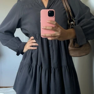 Mörkgrå volangklänning med knappar - Säljer en mörkgrå klänning med volanger och knappar framtill. Klänningen har lång ärm med manschett och en lös passform. Perfekt för dig som gillar en avslappnad men ändå stilren look.