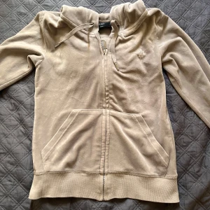 Beige sett från Juicy Couture - Säljer ett beige sett från Juicy Couture med dragkedja framtill och huva med snörning. Tröjan har långa ärmar, ribbade muddar och två stora fickor framtill. Perfekt för en avslappnad stil. Byxorna har små fläckar som jag inte testat behandla. Skriv för fler bilder, tryck inte köp nu skriv till mig innan💞pris kan diskuteras! Går även att köpa separat!