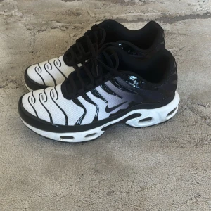 Nike Air Max Plus vita och svarta sneakers - Säljer ett par Nike Air Max Plus sneakers i vitt och svart med vågformade svarta linjer och svart sula. Skorna har snörning och den klassiska Nike-loggan på sidan. Perfekta för dig som gillar en sportig och modern stil.