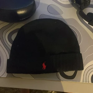 Svart mössa från Polo Ralph Lauren - Svart ribbstickad mössa från Polo Ralph Lauren med röd broderad logga framtill. Klassisk och stilren design som passar perfekt till kyliga dagar.