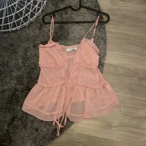 Rosa volangtop från Design by Amor - Säljer en söt och luftig rosa volangtop från Design by Amor. Toppen har tunna axelband, volangdetaljer och knytning framtill. Perfekt för varma dagar. Aldrig andvänd helt nytt skick❤️💕nypris 350 säljer för 300❤️