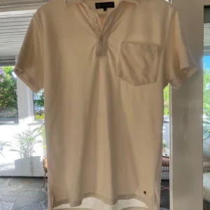 Vit piké t-shirt från Lagency - Säljer en stilren vit piké t-shirt från Lagency med klassisk krage, knappar vid halsen och en bröstficka. T-shirten har korta ärmar och är tillverkad i mjukt bomullsmaterial. Perfekt för en clean och avslappnad look.