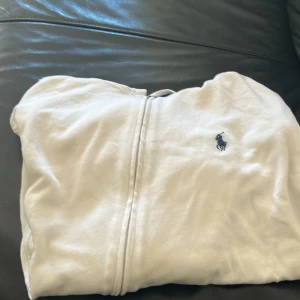 Vit hoodie från Polo Ralph Lauren - Säljer en klassisk vit hoodie från Polo Ralph Lauren med dragkedja och broderad logga på bröstet. Tröjan har huva och är tillverkad i mjukt bomullsmaterial. Perfekt för en avslappnad och stilren look. Scick 7/10 då den har lite fläckar som nog kan gå bort i en tvätt priset är ej hugget i sten