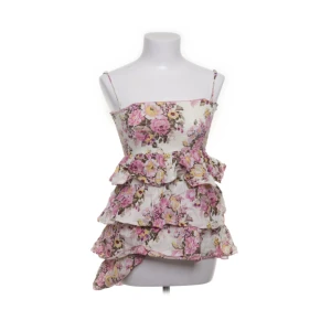 Blommig volangtopp från Poetry Clothing - Supersöt blommig topp från Poetry Clothing med volanger i flera lager och smala axelband. Toppen är i bomull och har ett romantiskt mönster i rosa, lila och gult på vit botten