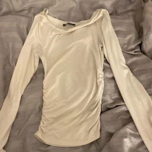 Vit långärmad topp från Gina Tricot - Säljer en vit långärmad topp från Gina Tricot i storlek XS. Toppen har en lätt asymmetrisk halsringning och rynkade detaljer längs ena sidan som ger en snygg passform. Perfekt att styla till olika outfits!