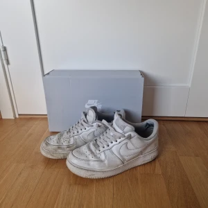 Vita Nike Air Force 1 sneakers - Klassiska vita Nike Air Force 1 sneakers med lågt skaft och perforerad tå. Skorna har snörning och är tillverkade i skinn med en platt sula. Perfekta för dig som gillar en tidlös och sportig stil.