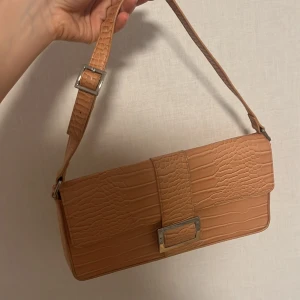 Väska  - Säljer en beige axelväska från Monki i krokopräglad syntet med justerbart axelband och silverfärgat spänne framtill. Den har bara använts två gånger 🫶 kontakta vid mer information 