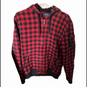 Polo Ralph lauren cardigan - Röd och svart rutig hoodie med dragkedja och huva från Polo Ralph Lauren. Jackan har ribbade muddar och fickor framtill samt broderad logga på bröstet. Perfekt nu när det är vår! Tveka inte på att höra av er ifall ni har några frågor.