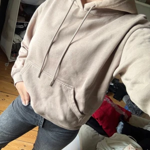 Beige hoodie med ficka framtill - Säljer en beige hoodie med huva och dragsnören. Tröjan har en stor magficka och ribbade muddar vid ärmslut och nederkant. Perfekt för en avslappnad stil.