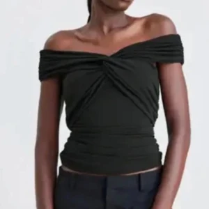 Svart offshoulder topp - Säljer en svart offshoulder topp med twistad detalj framtill. Toppen är figurnära och har en snygg drapering över bysten. Perfekt för att lyfta din outfit med en trendig touch.