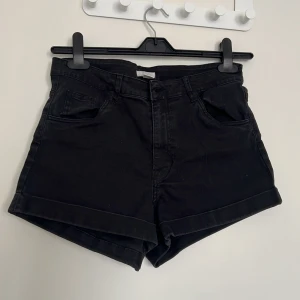 Svarta jeansshorts med upprullad kant - Svarta jeansshorts med upprullad kant och klassisk femficksdesign. Hög midja och stängs med knapp och dragkedja. Perfekta för varma dagar och enkla att matcha med olika toppar.