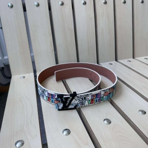 Louis Vuitton bälte med färgglatt mönster - Säljer ett Louis Vuitton bälte med färgglatt, grafiskt mönster och svart LV-spänne. Bältet är gjort i skinn och har tryckt logotyp. Perfekt för dig som vill sticka ut med accessoarer från ett lyxigt märke.