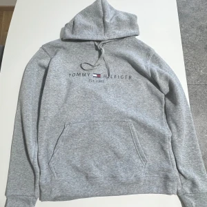 Grå hoodie från Tommy Hilfiger - Säljer en klassisk grå hoodie från Tommy Hilfiger med logotyp och texten 'Est. 1985' på bröstet. Tröjan har en justerbar huva med snören och ribbade muddar vid ärmslut och nederkant. Perfekt för en avslappnad stil. 