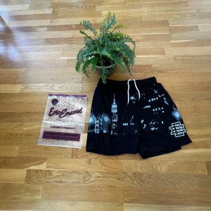 Eric Emanuel shorts - Säljer ett par svarta shorts från Eric Emanuel med vit stadssilhuett och logga på benet. Shortsen har snörning i midjan och är tillverkade i ett lätt, syntetiskt material. Perfekta för sommaren och har en normal passform. Jätte bra om man vill träna 