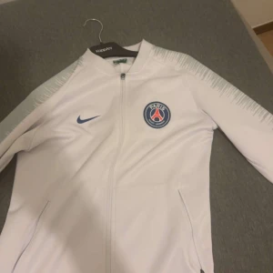 Vit Paris Saint-Germain - Säljer en vit träningsjacka från Nike med Paris Saint-Germain-logga på bröstet. Jackan har dragkedja framtill, långa ärmar med mönstrade detaljer och två fickor med dragkedja. Perfekt för fotbollsentusiasten! Storlek S passar även M