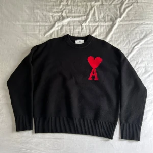 Svart stickad tröja från Ami Paris - Säljer en svart stickad tröja från Ami Paris med röd hjärt- och A-logga på bröstet. Tröjan har rund halsringning och ribbade muddar vid ärmslut och nederkant. Perfekt för dig som gillar stilrena plagg med en twist.