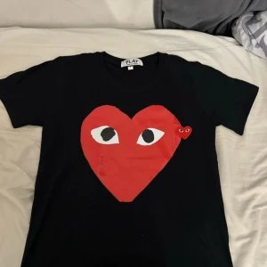 Svart t-shirt med rött hjärta från Comme des Garçons Play - Svart t-shirt från Comme des Garçons Play med stort rött hjärta och ögon på bröstet. Klassisk rund halsringning och korta ärmar. Perfekt statement-plagg för en trendig look.