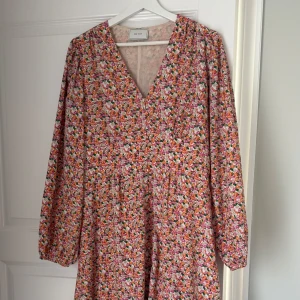 Blommig långklänning från Neo Noir - Säljer en färgglad långklänning från Neo Noir med blommigt mönster i orange, lila och grönt. Klänningen har v-ringning och långa ärmar, perfekt för vår och sommar. Materialet känns lätt och luftigt.