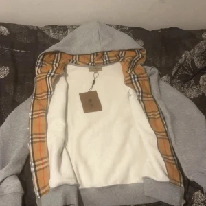 Grå hoodie med dragkedja från Burberry - Säljer en grå hoodie från Burberry med klassiskt rutigt foder i huvan. Tröjan har dragkedja framtill och ribbade muddar vid ärmslut och nederkant. Perfekt för dig som gillar stilrena detaljer och exklusiva märken.
