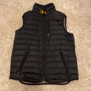 Svart väst från Parajumpers - Snygg svart väst från Parajumpers med hög krage och dragkedja framtill. Västen har två snedställda fickor med dragkedja och en insida i grått med säkerhetsficka. Perfekt för lager-på-lager under kyliga dagar. Äkta och inga skador på västen. Priset är inte hugget i sten så det är bara o skicka bud annars.