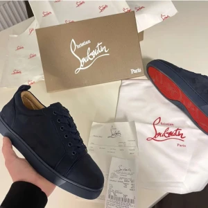 Mörkblå sneakers från Christian Louboutin - Säljer ett par mörkblå sneakers från Christian Louboutin med klassisk röd sula. Skorna har rund tå, platt sula och är tillverkade i mocka. Snygg broderad logga på baksidan och snörning.