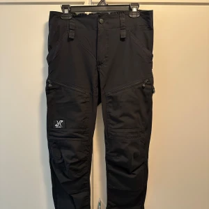 Svarta friluftsbyxor från RVRC - Svarta GPx Pro Pants från RVRC med flera dragkedjeförsedda fickor och förstärkta partier. Byxorna har elastisk midja och detaljerade sömmar för extra rörlighet. Perfekta för utomhusaktiviteter och äventyr.