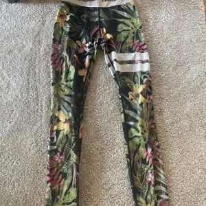 Färgglada blommiga träningstights - Säljer ett par färgglada träningstights med blommigt mönster och tropiska blad. Ifrån stringer. Tightsen har hög midja och vita detaljer på ena benet. Perfekta för gymmet eller löpning.