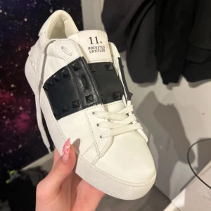 Valentino Rockstud Untitled sneakers med svarta nitar - Lägger upp igen pågrund av att skorna var för små för säljaren. Säljer ett par vita Valentino Rockstud Untitled sneakers med bred svart rem och svarta nitar över vristen. Skorna har rund tå, snörning och är tillverkade i skinn. Perfekta för dig som gillar lyxiga detaljer och stilren design. 39 men känns som 38/37,5⚠️ INGA RETURER⚠️