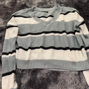 Randig stickad tröja med v-ringning - Säljer en stickad tröja med breda ränder i grått, vitt och svart. Tröjan har v-ringad hals och långa ärmar. Perfekt för dig som gillar en avslappnad men snygg stil.