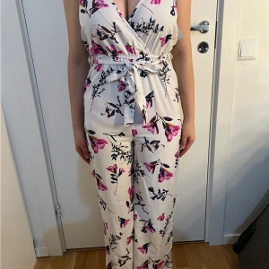 Vit blommig omlott jumpsuit med knytband - Säljer en vit jumpsuit med rosa och svarta blommor. Omlott överdel med v-ringning och knytband i midjan. Långa byxben och ärmlös design, perfekt för varmare dagar.