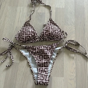Brunvit mönstrad bikini med knyt - Säljer en snygg bikini i brun och vit med grafiskt mönster. Både överdel och underdel har knytband i sidorna och nacken. Tillverkad i mjuk polyesterblandning med elastan för skön passform. Perfekt för stranden eller poolen! Storlek M, aldrig använd