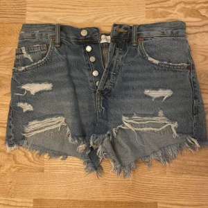 Blå jeansshorts från Zara - Säljer ett par blå jeansshorts från Zara med slitna detaljer och fransig kant. Shortsen har knappar framtill och klassiska fickor både fram och bak. Perfekta för sommaren!❤️