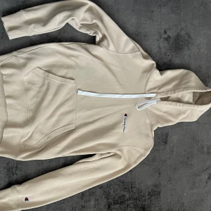 Beige hoodie från Champion - Säljer en beige hoodie från Champion med vit dragkedja och klassisk logga på bröstet. Tröjan har magficka och långa ärmar, perfekt för en avslappnad stil. Supermjuk och skön att ha på sig!