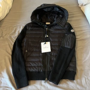 Svart Moncler cardigan - Snygg svart jacka från Moncler med quiltad front och huva. Ärmar och rygg är stickade för extra komfort och stil. Jackan har dragkedja, ribbade muddar och en praktisk ficka med dragkedja på ärmen. Perfekt för kyliga dagar. Passar S också!