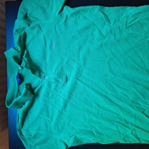 Grön pikétröja från GANT - Snygg grön pikétröja från GANT i 100% bomull. Klassisk krage med knappar framtill och liten broderad logga på bröstet. Perfekt för en stilren och avslappnad look.