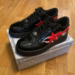 Svarta BAPE sneakers med röd stjärna och hajdetalj - Säljer ett par svarta BAPE sneakers i glansigt syntetmaterial med röd stjärna och hajmotiv på sidan. Skorna har snörning, platt sula och färgglada bokstäver bak. Kommer med originalkartong och BAPE-tag. Knappt använda. Pris kan diskuteras