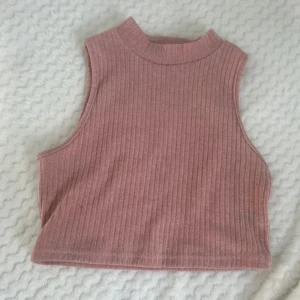 Rosa ribbad croppad linnetopp - Säljer en rosa croppad linnetopp med ribbad struktur och hög hals. Toppen är ärmlös och har en mjuk, stretchig känsla som passar perfekt till sommaren.
